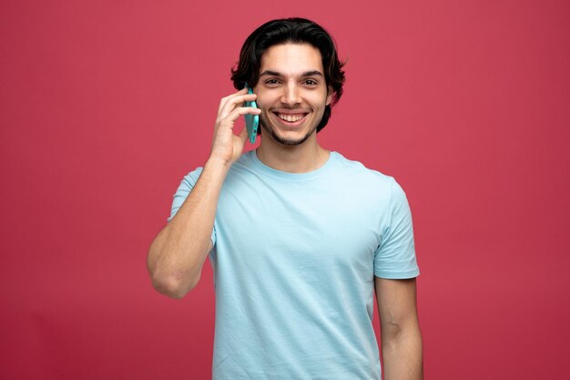 smiling young handsome man looking at camera talking on phone isolated on red background