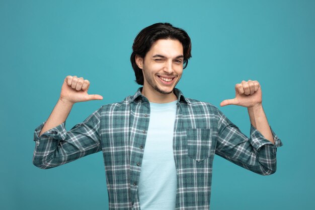 smiling young handsome man looking at camera winking pointing at himself isolated on blue background