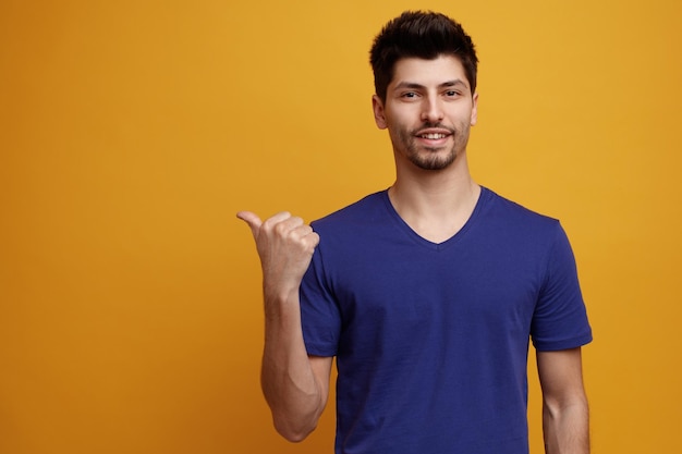 Smiling young handsome man pointing to side looking at camera on yellow background with copy space