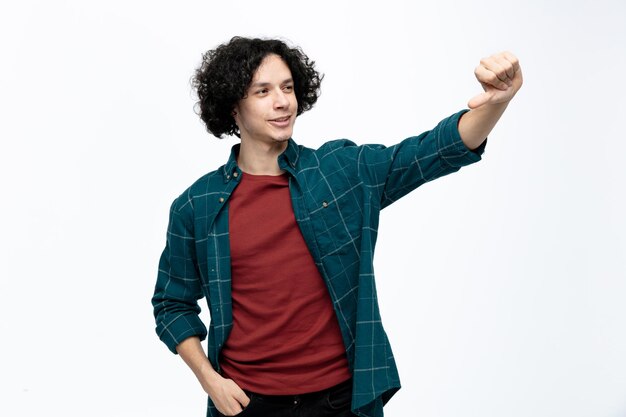 Smiling young handsome man pointing to side looking at his hand keeping another hand in pocket isolated on white background