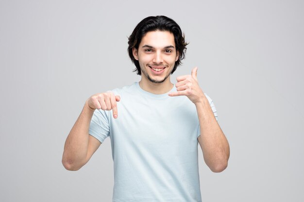 Smiling young handsome man showing call gesture pointing down while looking at camera isolated on white background