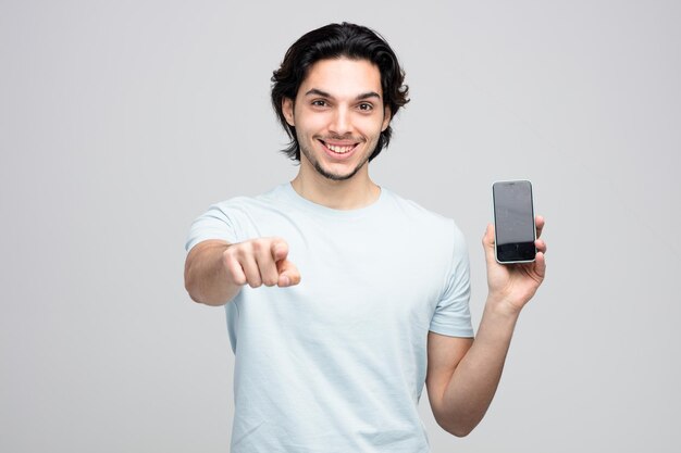 улыбающийся молодой красивый мужчина показывает мобильный телефон, глядя и указывая на камеру, изолированную на белом фоне
