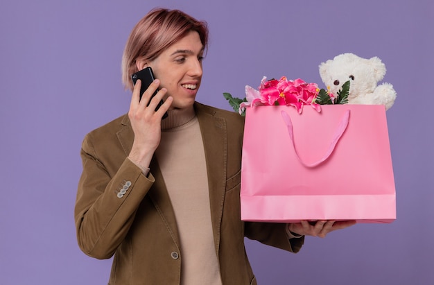 웃고 있는 젊고 잘생긴 남자는 전화 통화를 하고 꽃과 테디베어가 든 분홍색 선물 가방을 보고 있다