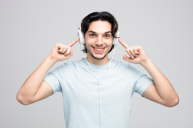 Foto gratuita sorridente giovane bell'uomo che indossa le cuffie che punta il dito su di loro guardando la fotocamera isolata su sfondo bianco