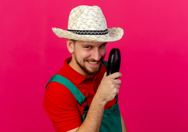 Foto gratuita sorridente giovane giardiniere slavo bello in uniforme e cappello che tiene alla ricerca di melanzane