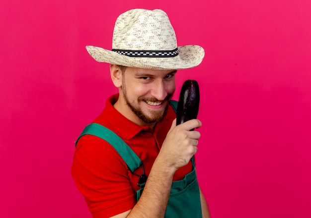 Smiling young handsome slavic gardener in uniform and hat holding aubergine looking 