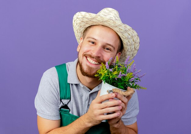 コピースペースと紫色の壁に孤立して見える植木鉢を保持している制服と帽子の若いハンサムなスラブの庭師の笑顔