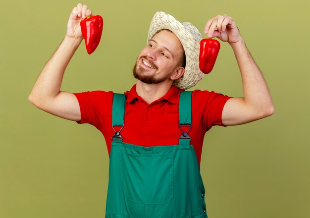 Улыбающийся молодой красивый славянский садовник в униформе и шляпе держит и смотрит на перец