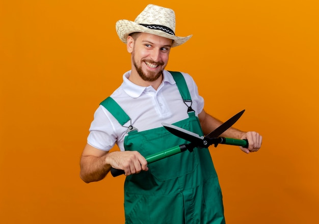 Smiling young handsome slavic gardener in uniform and hat holding pruners looking at side isolated