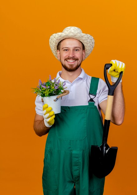 オレンジ色の壁に孤立して見えるスペードと植木鉢を保持している帽子と園芸用手袋を身に着けている制服を着た若いハンサムなスラブの庭師の笑顔