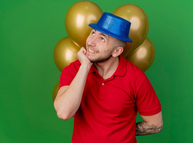 Smiling young handsome slavic party guy wearing party hat holding balloons behind back putting hand on chin looking up isolated on green background with copy space