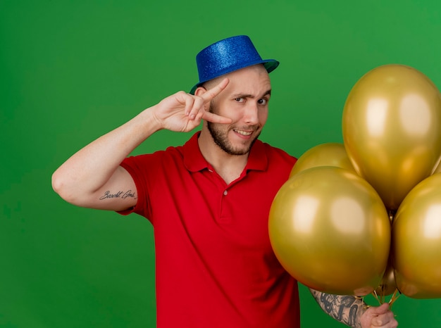 Smiling young handsome slavic party guy wearing party hat holding balloons doing peace sign looking at camera isolated on green background