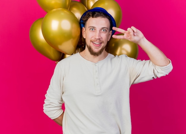 Free photo smiling young handsome slavic party guy wearing party hat holding balloons behind his back doing peace sign looking at camera isolated on crimson background