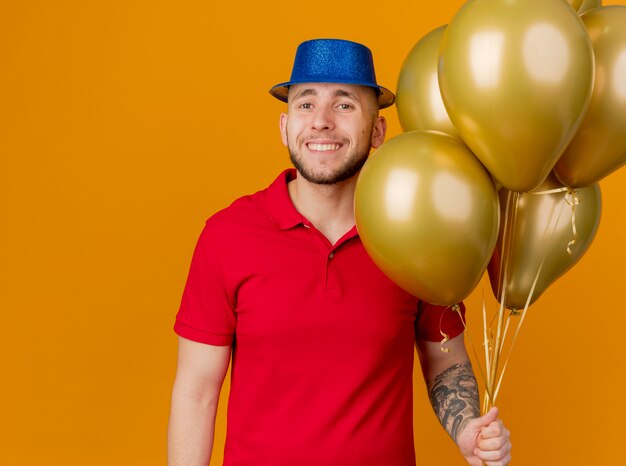 Улыбающийся молодой красивый славянский партийный парень в партийной шляпе держит воздушные шары, глядя в камеру, изолированную на оранжевом фоне с копией пространства