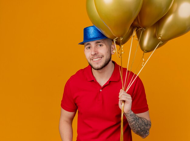 Smiling young handsome slavic party guy wearing party hat holding balloons looking at camera isolated on orange background with copy space