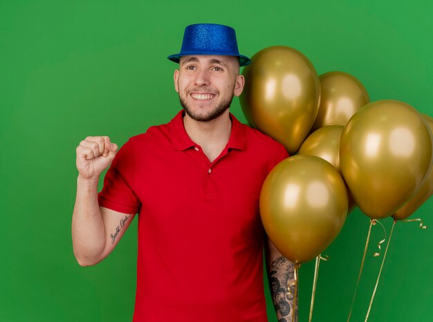 Smiling young handsome slavic party guy wearing party hat holding balloons looking at side doing yes gesture isolated on green background