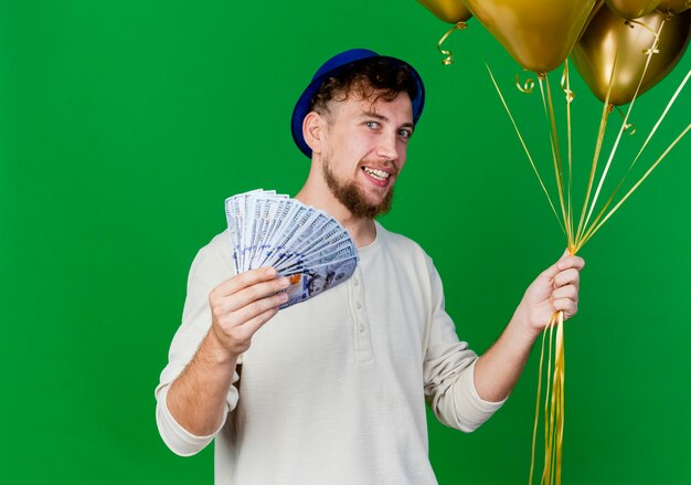 복사 공간이 녹색 배경에 고립 된 카메라를보고 풍선과 돈을 들고 파티 모자를 쓰고 젊은 잘 생긴 슬라브 파티 남자 미소