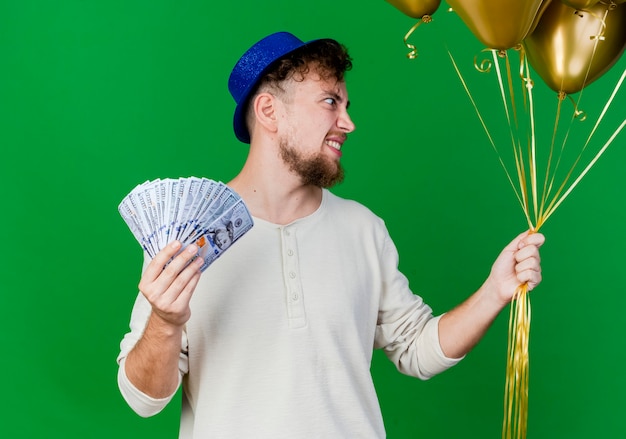 Smiling young handsome slavic party guy wearing party hat holding balloons and money looking at side isolated on green background