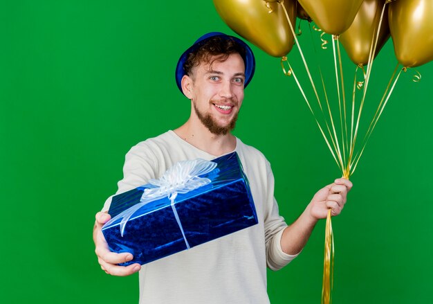 Smiling young handsome slavic party guy wearing party hat holding balloons and stretching out gift box towards camera isolated on green background with copy space