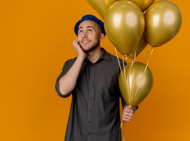 Smiling young handsome slavic party guy wearing party hat holding balloons touching face looking up isolated on orange background with copy space