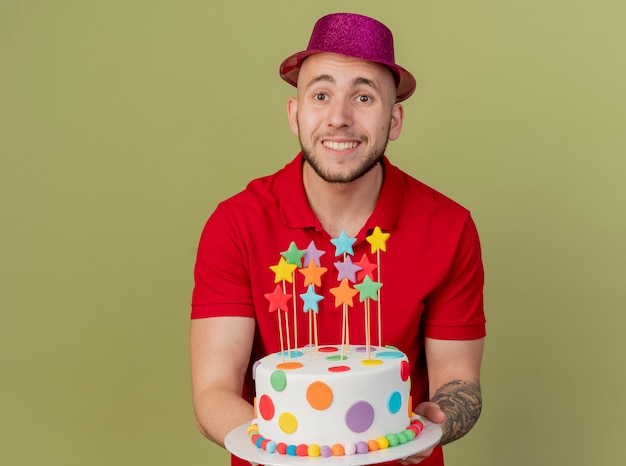 Foto gratuita sorridente giovane ragazzo bello partito slavo indossando il cappello del partito che tiene la torta di compleanno che guarda l'obbiettivo isolato su sfondo verde oliva con lo spazio della copia