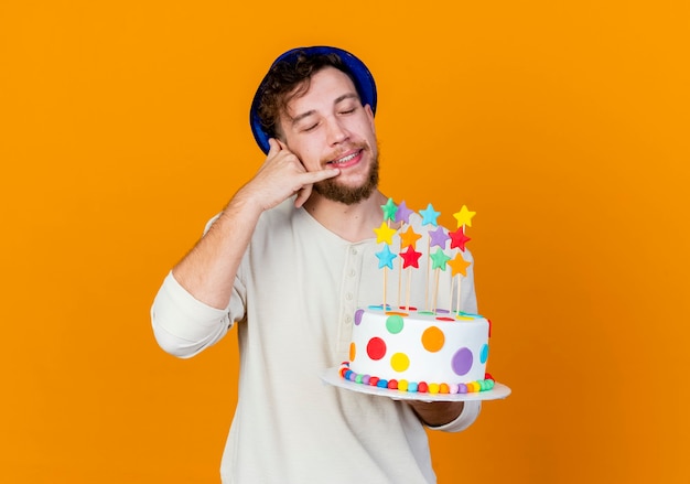 Smiling young handsome slavic party guy wearing party hat holding birthday cake with stars doing call gesture with closed eyes isolated on orange background with copy space