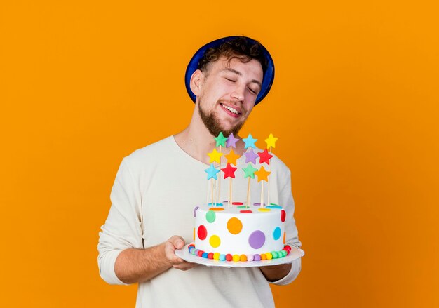 コピースペースとオレンジ色の背景に分離された星とバースデーケーキを保持しているパーティー帽子をかぶって若いハンサムなスラブ党の男を笑顔