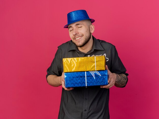 Smiling young handsome slavic party guy wearing party hat holding gift packs with closed eyes isolated on crimson background with copy space