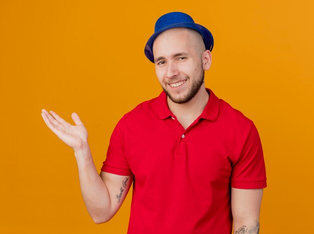 Smiling young handsome slavic party guy wearing party hat looking at camera showing empty hand isolated on orange background