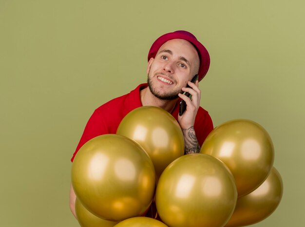 Smiling young handsome slavic party guy wearing party hat standing behind balloons talking on phone looking at side isolated on olive green background with copy space