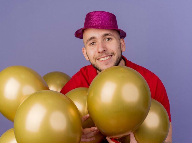 Smiling young handsome slavic party guy wearing party hat standing among balloons touching them looking at camera isolated on purple background