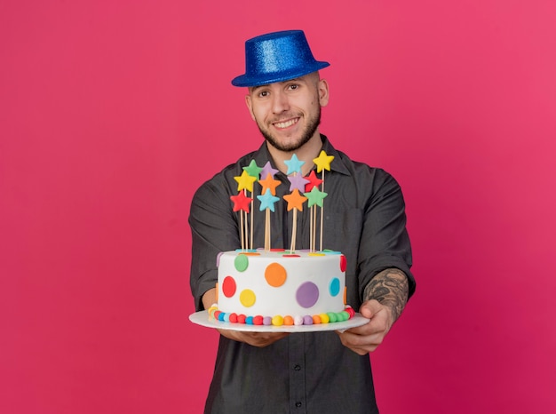 Free photo smiling young handsome slavic party guy wearing party hat stretching out birthday cake with stars towards camera looking at camera isolated on crimson background with copy space
