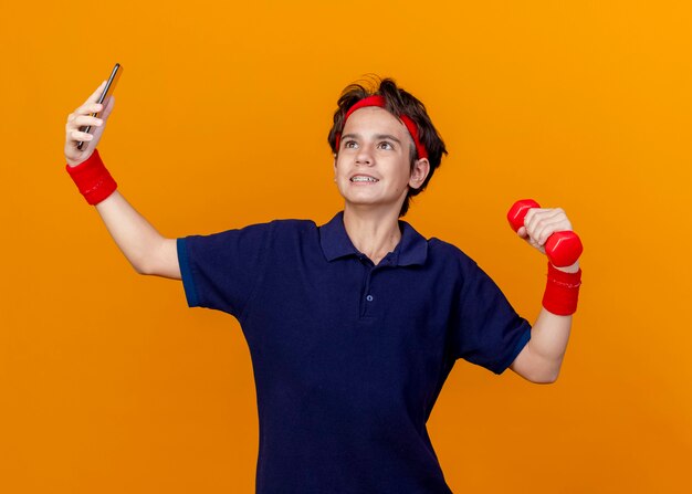 Smiling young handsome sporty boy wearing headband and wristbands with dental braces holding dumbbell looking up taking selfie isolated on orange wall