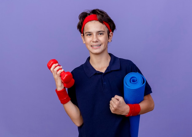 Smiling young handsome sporty boy wearing headband and wristbands with dental braces holding dumbbells and yoga mat  isolated on purple wall with copy space