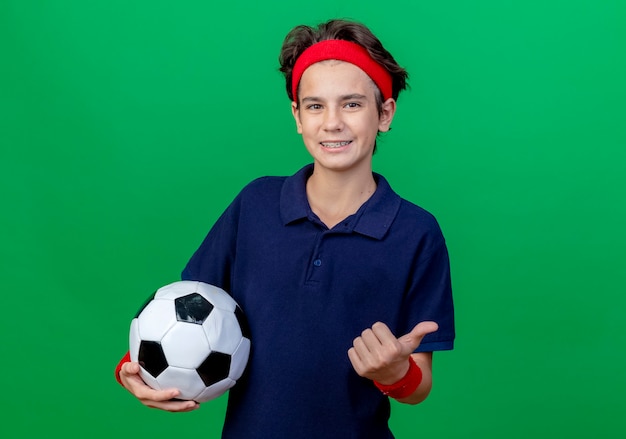 Smiling young handsome sporty boy wearing headband and wristbands with dental braces holding soccer ball showing thumb up looking at camera isolated on green background with copy space