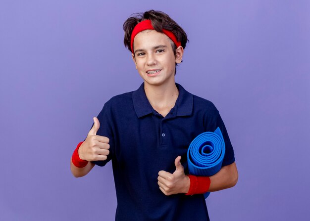 Улыбающийся молодой красивый спортивный мальчик с головной повязкой и браслетами с зубными скобами, держащий коврик для йоги, показывает палец вверх, изолированный на фиолетовой стене с копией пространства