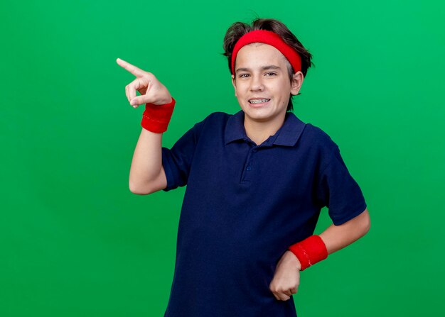 Smiling young handsome sporty boy wearing headband and wristbands with dental braces keeping hand on waist looking at front pointing at side isolated on green wall with copy space
