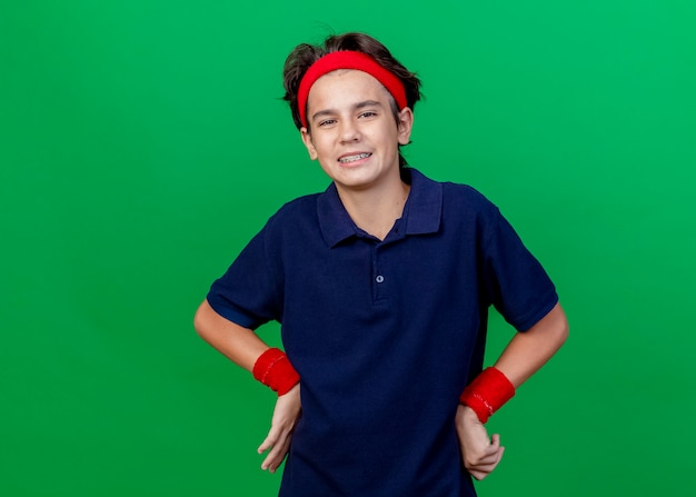 Smiling young handsome sporty boy wearing headband and wristbands with dental braces keeping hands on waist looking at camera isolated on green background with copy space