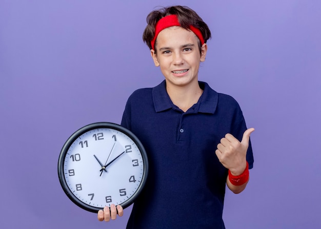 Foto gratuita sorridente giovane bel ragazzo sportivo che indossa la fascia e braccialetti con bretelle dentali guardando la fotocamera tenendo l'orologio che mostra il pollice in alto isolato su sfondo viola con spazio di copia