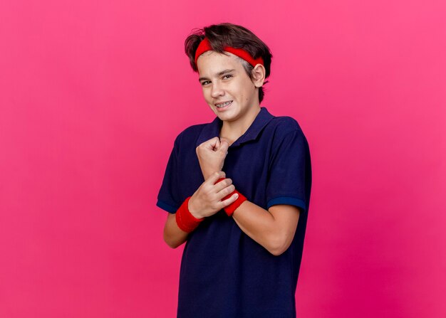 Smiling young handsome sporty boy wearing headband and wristbands with dental braces looking at camera holding wrist isolated on crimson background with copy space