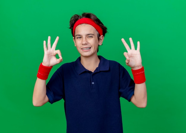 Smiling young handsome sporty boy wearing headband and wristbands with dental braces looking at front doing ok sign isolated on green wall