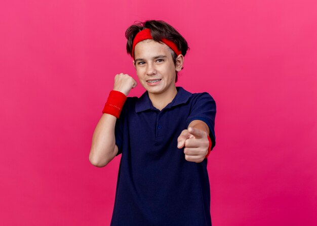 Smiling young handsome sporty boy wearing headband and wristbands with dental braces looking and pointing at camera clenching fist isolated on crimson background with copy space