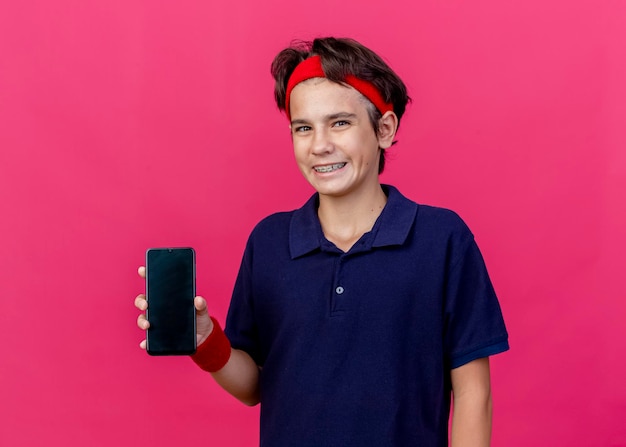 Sorridente giovane ragazzo sportivo bello che indossa la fascia e braccialetti con le parentesi graffe dentali che mostra il telefono cellulare guardando la parte anteriore isolata sulla parete rosa