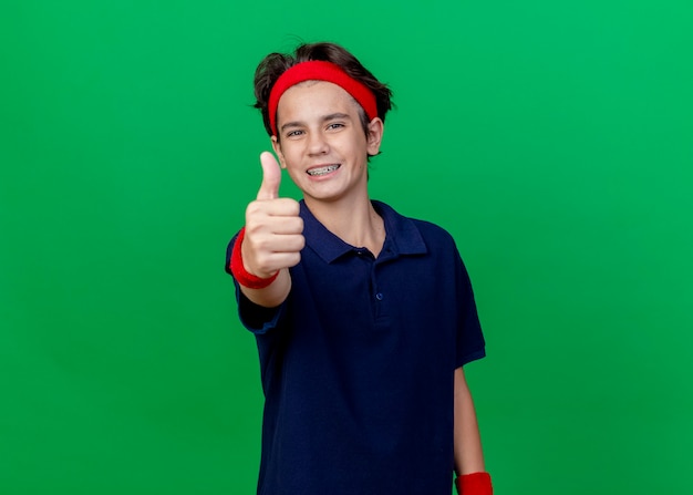 Smiling young handsome sporty boy wearing headband and wristbands with dental braces  showing thumb up isolated on green wall with copy space