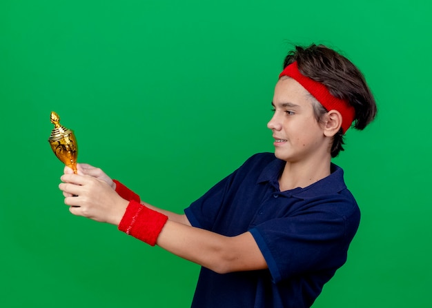 Smiling young handsome sporty boy wearing headband and wristbands with dental braces standing in profile view holding and looking at winner cup isolated on green wall