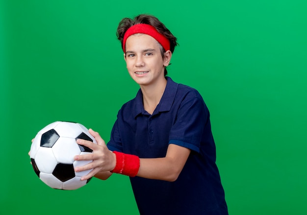 Sorridente giovane ragazzo sportivo bello che indossa la fascia e braccialetti con bretelle dentali che allunga il pallone da calcio isolato sulla parete verde con lo spazio della copia