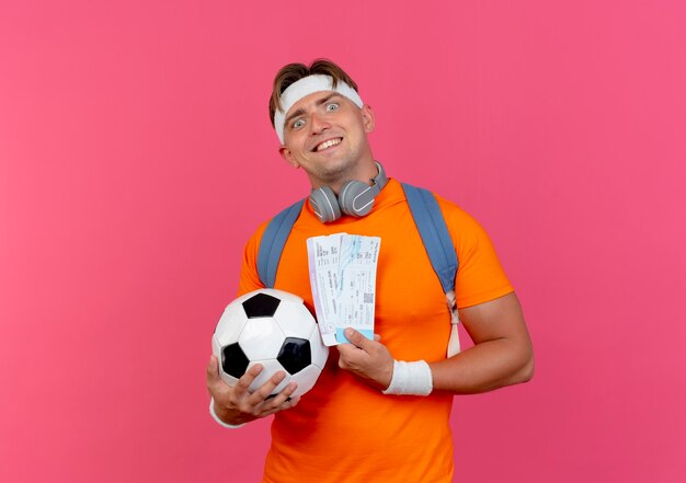 Smiling young handsome sporty man wearing headband and wristbands and back bag with headphones on neck holding airplane tickets and soccer ball isolated on pink wall