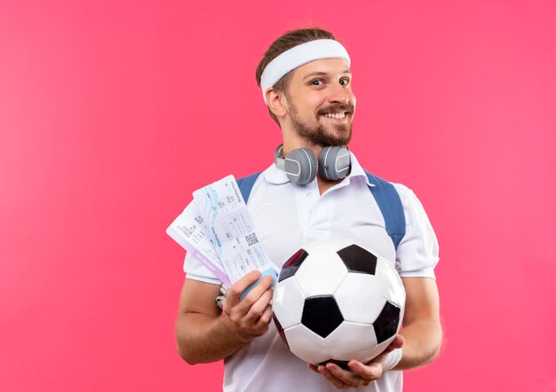 Smiling young handsome sporty man wearing headband and wristbands and back bag with headphones on neck holding soccer ball and airplane tickets isolated on pink space 