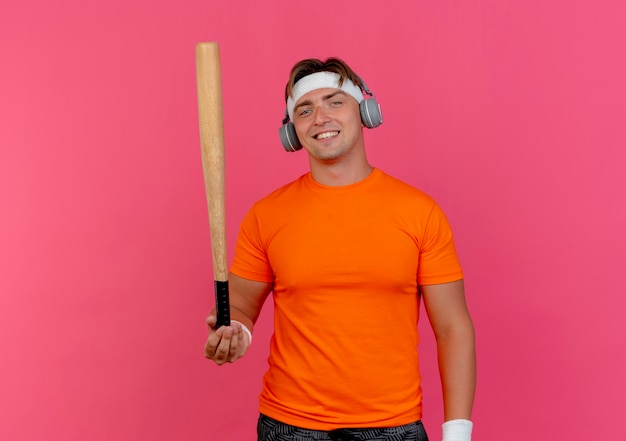 Smiling young handsome sporty man wearing headband and wristbands and headphones holding baseball bat isolated on pink wall