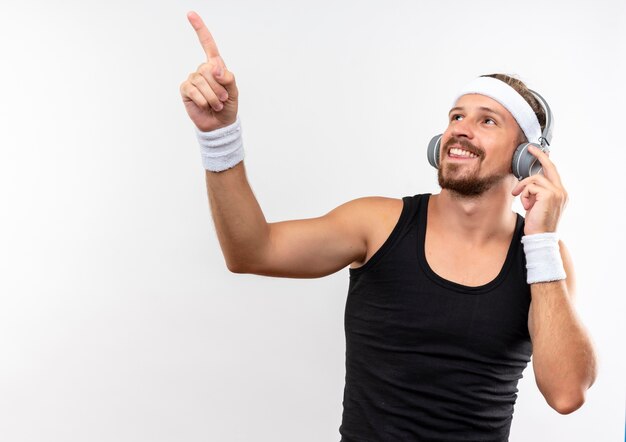Smiling young handsome sporty man wearing headband and wristbands and headphones looking and pointing up with one hand on headphone isolated on white space 